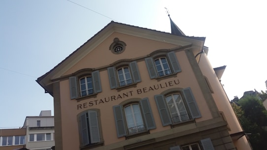 A European-style building with beige walls and multiple windows featuring blue shutters. The text 'Restaurant Beaulieu' is prominently displayed. The building has a triangular roof with an architectural detail near the top, possibly a small circular window or decoration. This establishment is situated in an area with additional modern buildings visible in the background.