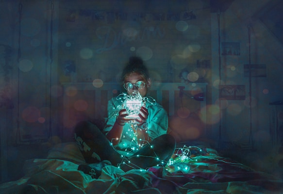 A person sits on a bed in a dimly lit room, holding a string of glowing fairy lights, which casts a soft, ambient glow. The atmosphere is cozy and dreamlike with soft bokeh lights scattered throughout the scene.