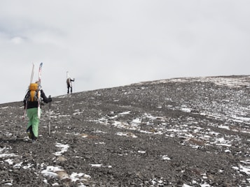 Two hikers are climbing a rocky, snow-dusted mountain slope. They are wearing backpacks and carrying skis, indicating they are on a skiing expedition. The sky is cloudy and overcast, creating a stark, isolated atmosphere.