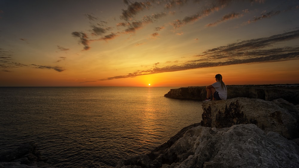woman sitting on top of the cliff looking at the horizon of sunrise near body of water