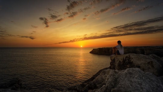 woman sitting on top of the cliff looking at the horizon of sunrise near body of water in Menorca Spain