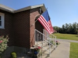 A brick building with a set of white metal railings leading up to a small entrance with a cross on top. An American flag is displayed prominently on a pole attached to the building. There are various potted plants and flowers around the base of the railings. In the background, a well-maintained lawn and a road can be seen under a clear blue sky.
