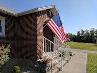 A brick building with a set of white metal railings leading up to a small entrance with a cross on top. An American flag is displayed prominently on a pole attached to the building. There are various potted plants and flowers around the base of the railings. In the background, a well-maintained lawn and a road can be seen under a clear blue sky.