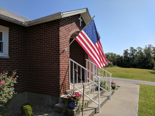 A brick building with a set of white metal railings leading up to a small entrance with a cross on top. An American flag is displayed prominently on a pole attached to the building. There are various potted plants and flowers around the base of the railings. In the background, a well-maintained lawn and a road can be seen under a clear blue sky.