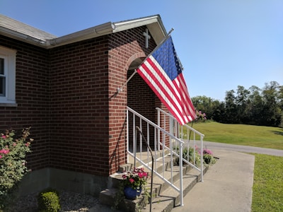 A brick building with a set of white metal railings leading up to a small entrance with a cross on top. An American flag is displayed prominently on a pole attached to the building. There are various potted plants and flowers around the base of the railings. In the background, a well-maintained lawn and a road can be seen under a clear blue sky.