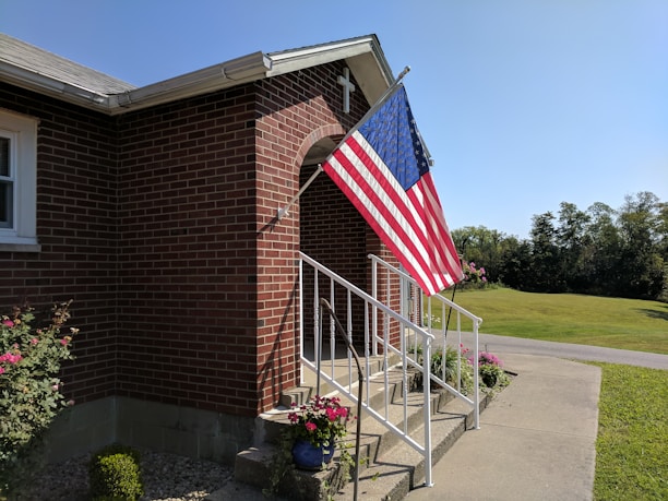 A brick building with a set of white metal railings leading up to a small entrance with a cross on top. An American flag is displayed prominently on a pole attached to the building. There are various potted plants and flowers around the base of the railings. In the background, a well-maintained lawn and a road can be seen under a clear blue sky.