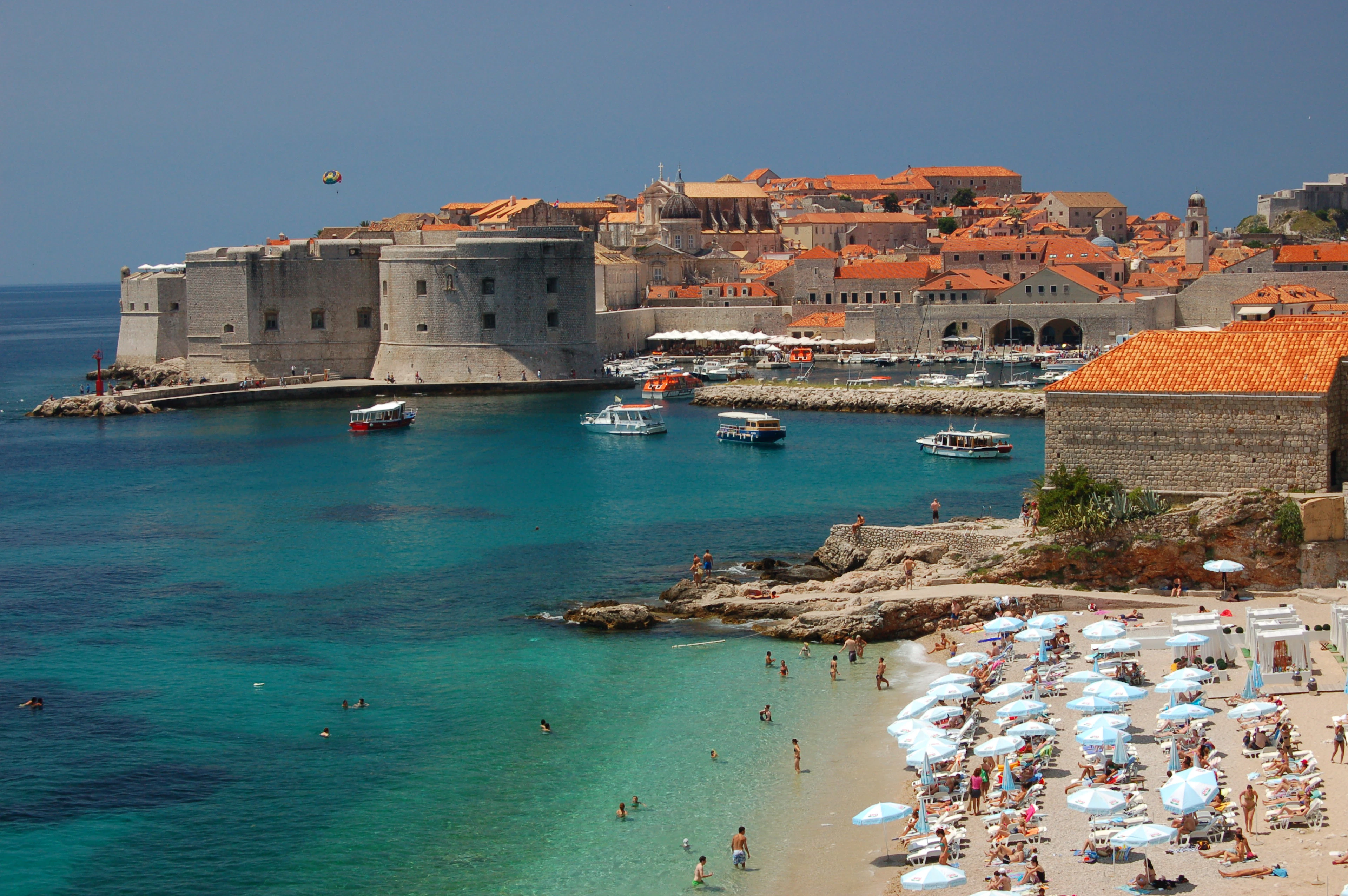 Banje Beach in Dubrovnik on a bright summer day, rows of white parasols and bathers in turquoise shallows, with St John's Fortress, the Old Port and the terracotta roofs of the Old Town beyond.
