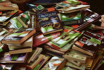 A large pile of various books, mostly paperbacks, in different colors and sizes. Many books have clearance sale stickers on them. The books are stacked haphazardly, overlapping each other. Some titles and covers are clearly visible while others are partially obscured.