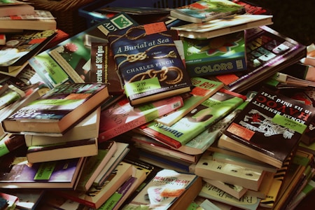 A large pile of various books, mostly paperbacks, in different colors and sizes. Many books have clearance sale stickers on them. The books are stacked haphazardly, overlapping each other. Some titles and covers are clearly visible while others are partially obscured.
