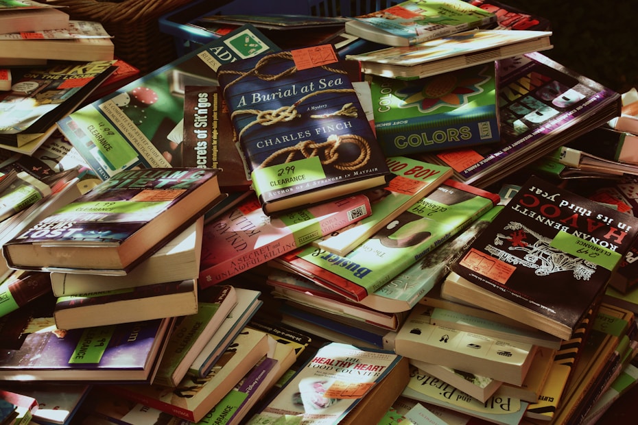 A large pile of various books, mostly paperbacks, in different colors and sizes. Many books have clearance sale stickers on them. The books are stacked haphazardly, overlapping each other. Some titles and covers are clearly visible while others are partially obscured.