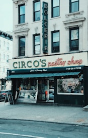 A storefront of a pastry shop with vintage-style signage and an overarching canopy, featuring a large display window and double doors. The building has classic architecture with detailed moldings around the windows. Above, additional signage reads 'Dolceria' vertically.