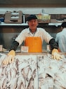 man wearing white polo shirt and brown apron standing near fish stands