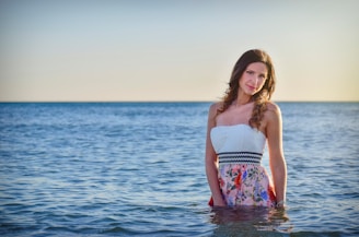 A woman stands in the sea wearing a white strapless dress with colorful floral patterns. The tranquil ocean stretches to the horizon under a clear sky, with the lighting suggesting a sunset or sunrise.