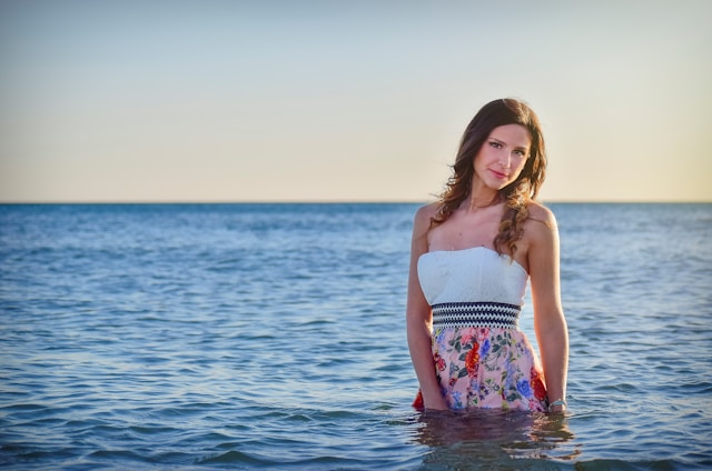 A woman stands in the sea wearing a white strapless dress with colorful floral patterns. The tranquil ocean stretches to the horizon under a clear sky, with the lighting suggesting a sunset or sunrise.