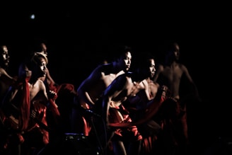 A group of shirtless individuals is captured in motion on a dimly lit stage, their bodies partially illuminated and wrapped in red fabric. The scene suggests a dynamic and intense performance, with the figures striking powerful and energetic poses.