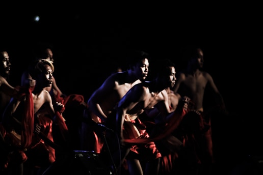 A group of shirtless individuals is captured in motion on a dimly lit stage, their bodies partially illuminated and wrapped in red fabric. The scene suggests a dynamic and intense performance, with the figures striking powerful and energetic poses.