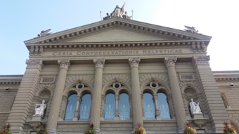 A grand neoclassical building facade featuring tall Corinthian columns and intricate architectural details. The structure is adorned with statues and has large arched windows. The inscription 'Curia Confoederationis Helveticae' is prominently displayed along the top.