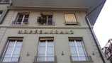 A building facade with rectangular windows, some of which are adorned with yellow and white striped awnings. The word 'HOPFENKRANZ' is prominently displayed in large, golden letters. Several potted plants are visible on the balcony.