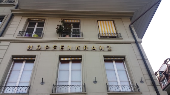 A building facade with rectangular windows, some of which are adorned with yellow and white striped awnings. The word 'HOPFENKRANZ' is prominently displayed in large, golden letters. Several potted plants are visible on the balcony.