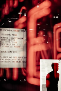 A silhouette of a person stands in front of a board with text, possibly a cinema listing. The background features blurred red neon lights creating an abstract and dynamic effect.