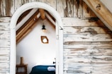 A cozy attic room with rustic wooden walls and a simple bed covered in dark blue bedding. A small wooden table with a rolled towel and wicker basket is visible. A geometric mirror hangs on the white wall beside a hanging lamp.