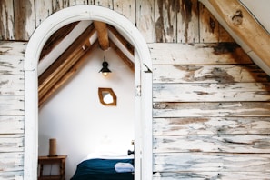 A cozy attic room with rustic wooden walls and a simple bed covered in dark blue bedding. A small wooden table with a rolled towel and wicker basket is visible. A geometric mirror hangs on the white wall beside a hanging lamp.