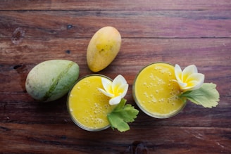 Two ripe mangoes and two glasses of mango smoothie are placed on a wooden table. Each smoothie glass is garnished with a white and yellow flower and a green leaf.