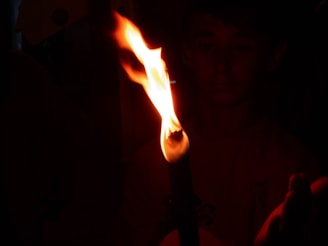 A torch with a bright, flickering flame illuminates the darkness, casting a warm glow. Parts of a person's face and hand are faintly visible in the background, highlighting the dim environment.