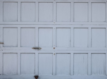 An old, weathered garage door with a pattern of rectangular panels. The panels have a white, slightly peeling paint finish. There are small metallic handles and fixtures visible on the surface.