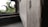 Close-up of a wooden window shutter with a blurred background showing part of a window frame and exterior greenery. The image focuses on the texture of the wood with visible grain patterns.