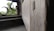 Close-up of a wooden window shutter with a blurred background showing part of a window frame and exterior greenery. The image focuses on the texture of the wood with visible grain patterns.