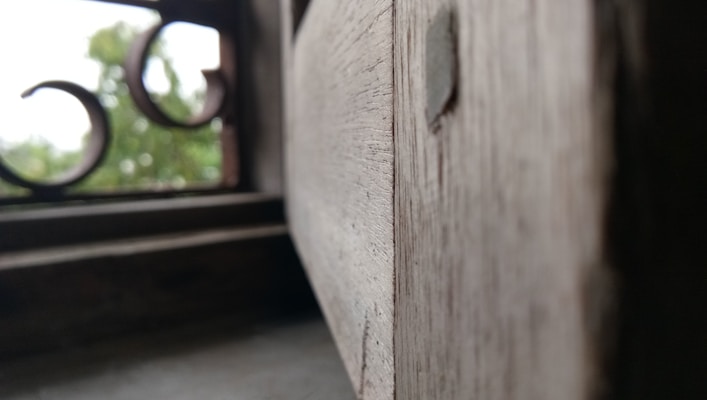 Close-up of a wooden window shutter with a blurred background showing part of a window frame and exterior greenery. The image focuses on the texture of the wood with visible grain patterns.