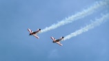 Two aerobatic airplanes are flying in formation against a clear blue sky. Each plane is emitting a trail of white smoke as they maneuver through the air. The airplanes have striking red and white designs on their wings and fuselages.