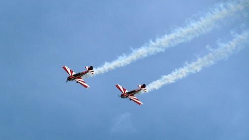 Two aerobatic airplanes are flying in formation against a clear blue sky. Each plane is emitting a trail of white smoke as they maneuver through the air. The airplanes have striking red and white designs on their wings and fuselages.