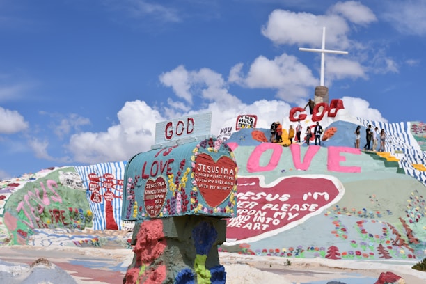 A colorful, artistic landscape with large words and religious messages painted on a hill. A group of people stands near the top beside a large cross. The phrases include 'God is Love' and 'Jesus I'm a Sinner.' The sky is clear with a few clouds.