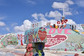 A colorful, artistic landscape with large words and religious messages painted on a hill. A group of people stands near the top beside a large cross. The phrases include 'God is Love' and 'Jesus I'm a Sinner.' The sky is clear with a few clouds.