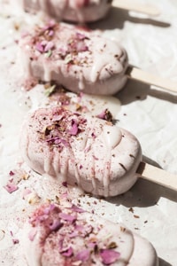 Five ice cream bars garnished with rose petals and dusted with a light powder are placed on a white surface. The ice cream is melting slightly, creating a creamy texture with the sunny warmth accentuating the delicate decorations.
