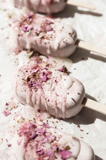 Five ice cream bars garnished with rose petals and dusted with a light powder are placed on a white surface. The ice cream is melting slightly, creating a creamy texture with the sunny warmth accentuating the delicate decorations.