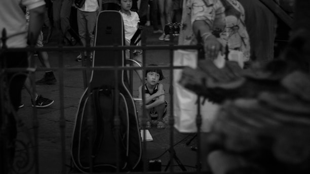 A child crouches on the ground, surrounded by a group of adults. The child appears contemplative, looking ahead with a focused gaze, while the adults are mostly not facing the camera. A large musical instrument case stands vertically nearby, suggesting a performance setting. The image is in black and white, creating a timeless, classic feel.