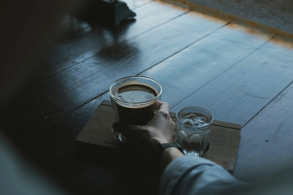 person holding brown liquid filled clear drinking glass beside clear liquid filled glass with ice cubes on brown wooden tray