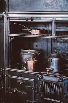A vintage cast iron stove is adorned with copper cookware, including a kettle and a pot, creating an atmosphere of old-world charm. The stove features intricate detailing and appears well-used, hinting at a historical or rustic setting.
