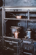 A vintage cast iron stove is adorned with copper cookware, including a kettle and a pot, creating an atmosphere of old-world charm. The stove features intricate detailing and appears well-used, hinting at a historical or rustic setting.