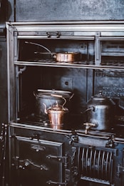A vintage cast iron stove is adorned with copper cookware, including a kettle and a pot, creating an atmosphere of old-world charm. The stove features intricate detailing and appears well-used, hinting at a historical or rustic setting.