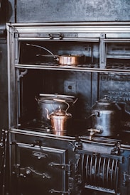 A vintage cast iron stove is adorned with copper cookware, including a kettle and a pot, creating an atmosphere of old-world charm. The stove features intricate detailing and appears well-used, hinting at a historical or rustic setting.