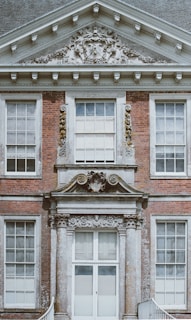 A detailed facade of a classic brick building featuring large, symmetrical windows and ornate stonework. The architectural embellishments include decorative reliefs and a coat of arms above the entrance.