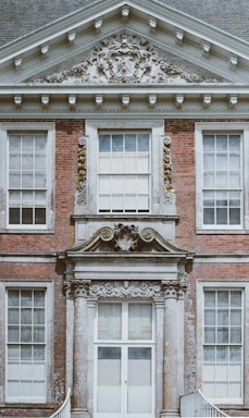 A detailed facade of a classic brick building featuring large, symmetrical windows and ornate stonework. The architectural embellishments include decorative reliefs and a coat of arms above the entrance.