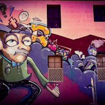 A colorful street art mural featuring a cartoonish character with an exaggerated facial expression, holding a heart in its hand. Surrounding the character are abstract shapes, including clouds, a speech bubble with a diamond, and various urban elements like a house and a can. The background includes a building wall with windows.
