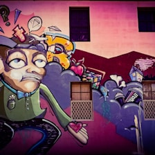 A colorful street art mural featuring a cartoonish character with an exaggerated facial expression, holding a heart in its hand. Surrounding the character are abstract shapes, including clouds, a speech bubble with a diamond, and various urban elements like a house and a can. The background includes a building wall with windows.