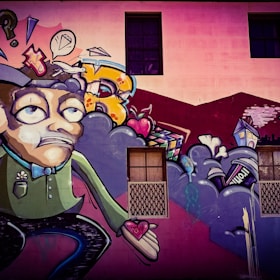 A colorful street art mural featuring a cartoonish character with an exaggerated facial expression, holding a heart in its hand. Surrounding the character are abstract shapes, including clouds, a speech bubble with a diamond, and various urban elements like a house and a can. The background includes a building wall with windows.