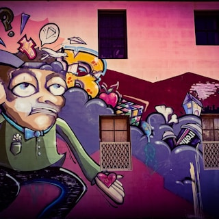 A colorful street art mural featuring a cartoonish character with an exaggerated facial expression, holding a heart in its hand. Surrounding the character are abstract shapes, including clouds, a speech bubble with a diamond, and various urban elements like a house and a can. The background includes a building wall with windows.